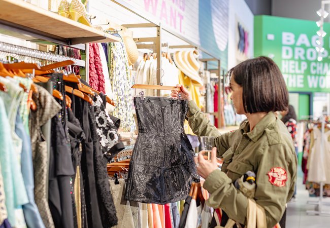 A woman with bobbed brown hair browses clothes at an Oxfam shop, looking at a short black dress.