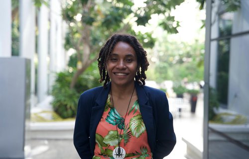 A woman from the Solomon Islands who is looking into the camera and smiling. She wears a pink top with tropical leaf pattern on it.