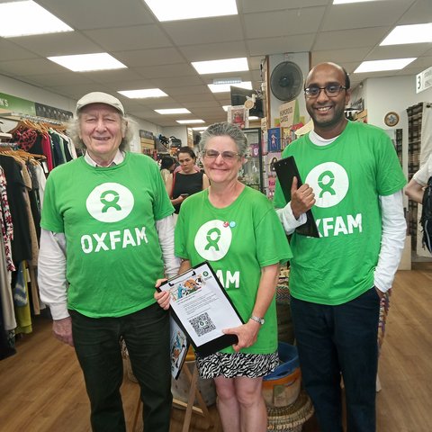 Three people wearing bright green Oxfam T-shirts stand inside a shop, holding clipboards and a tablet, with clothing racks and shelves visible in the background.