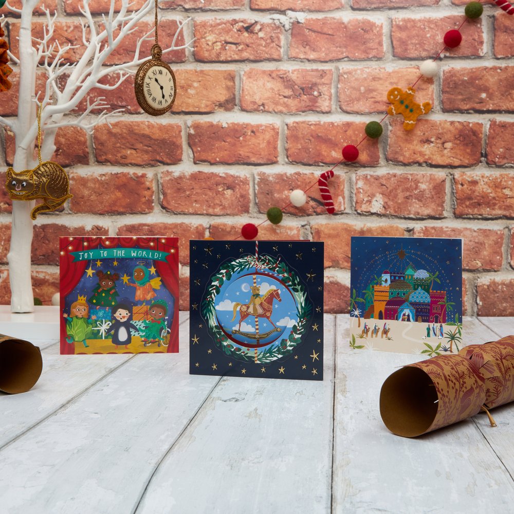 Three Christmas cards standing on a white hooden surface with the back drop of a brick wall and Christmas decorations in the back.