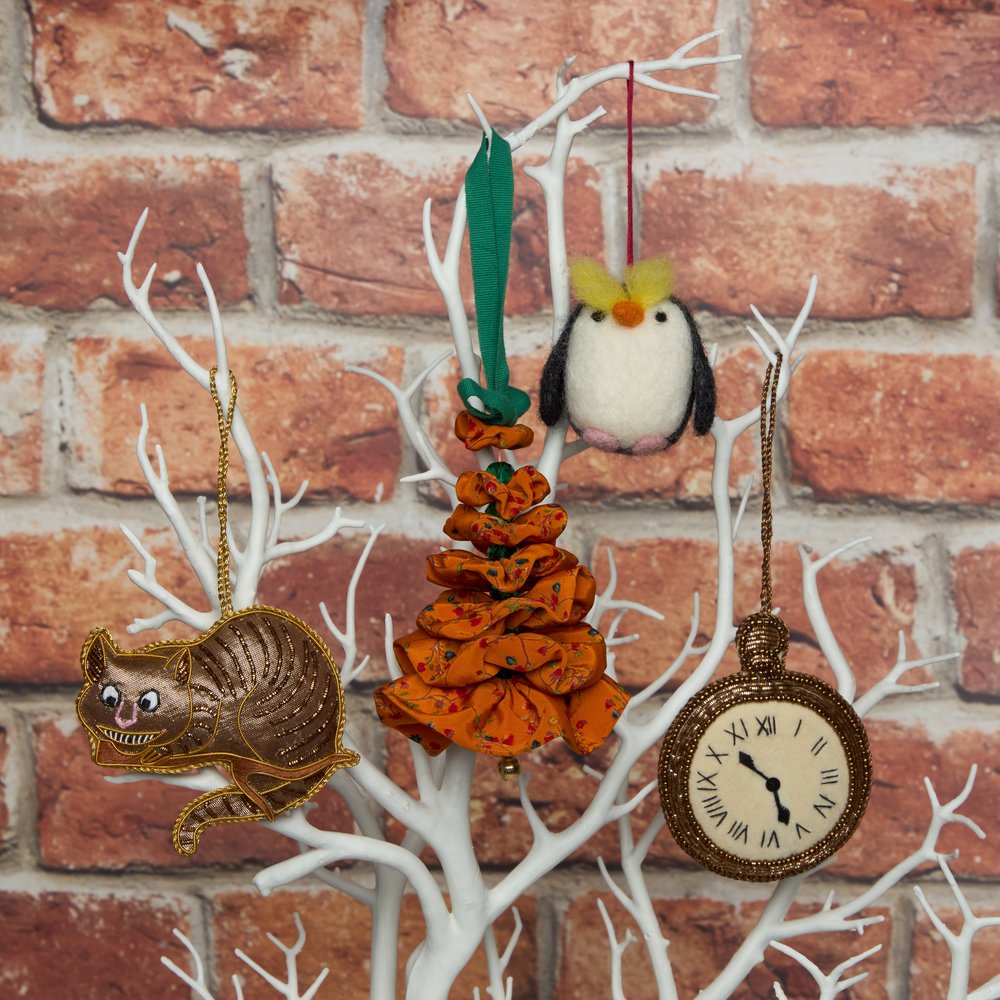 A decor tree with white branches behind a brick wall hanging Christmas tree decorations on, the decorations include of a orange Christmas tree centred in the middle  with a penguin, Cheshire cat and roman clock.