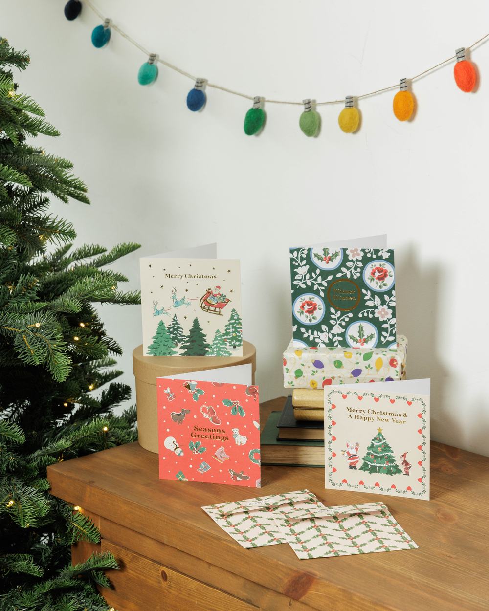 A collection of illustrated Christmas cards displayed on a wooden table beside a decorated Christmas tree and colourful festive garland.