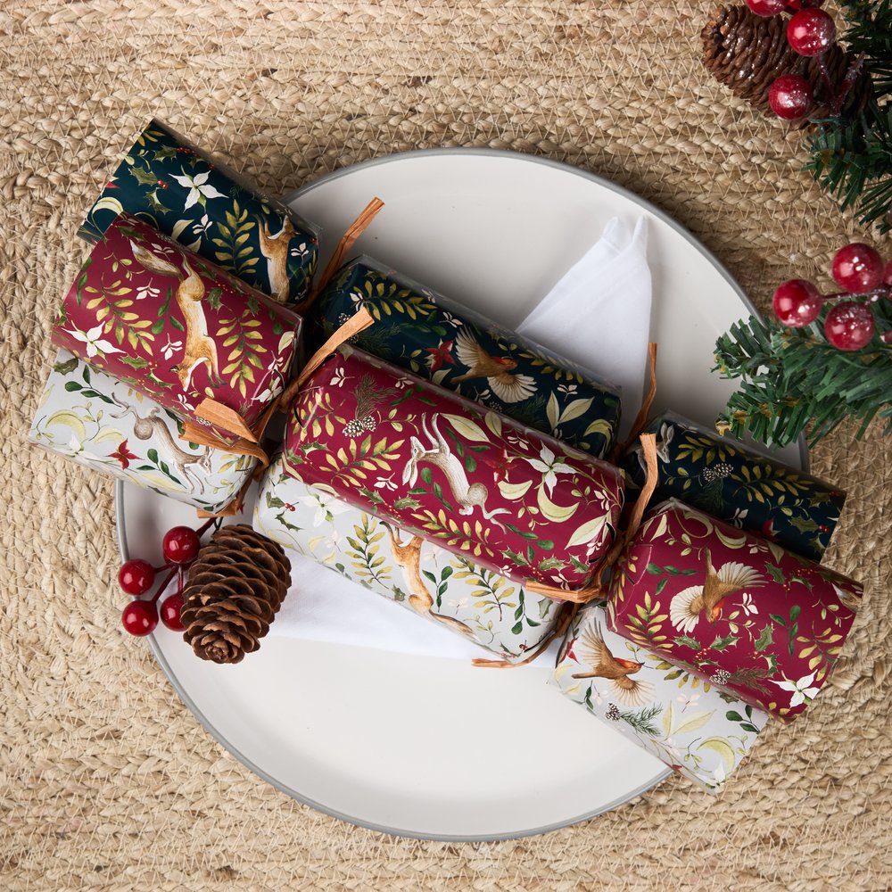 Three Christmas crackers with a woodland & animals pattern on on a green, red and grey crackers. They sit on a white plate with a pinecone, cranberries and features a branch of a Christmas tree.