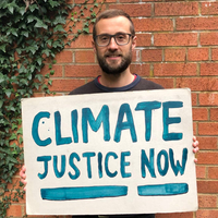 Oxfam Climate Campaigner Paul holds a sign outside his house that says 'climate justice now' in blue paint