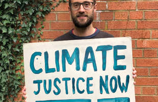 Oxfam Climate Campaigner Paul holds a sign outside his house that says 'climate justice now' in blue paint