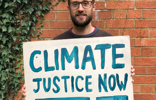 Oxfam Climate Campaigner Paul holds a sign outside his house that says 'climate justice now' in blue paint