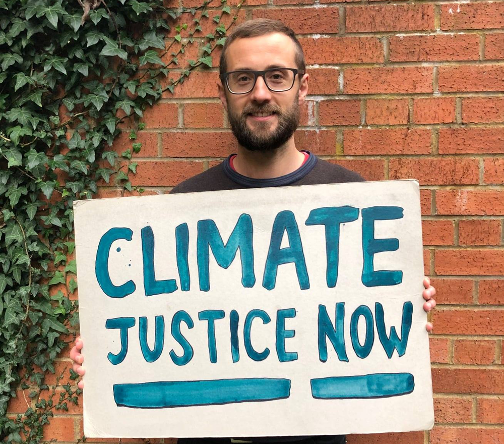 Oxfam Climate Campaigner Paul holds a sign outside his house that says 'climate justice now' in blue paint