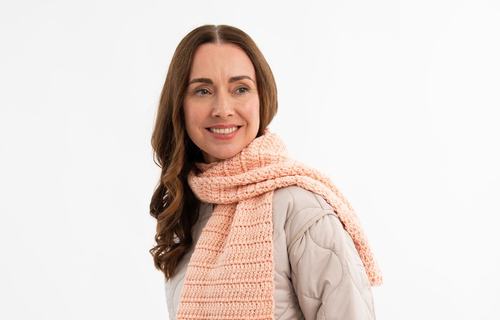 A person smiling at the camera wearing jeans, a beige puffer coat and a light pink crocheted scarf.
