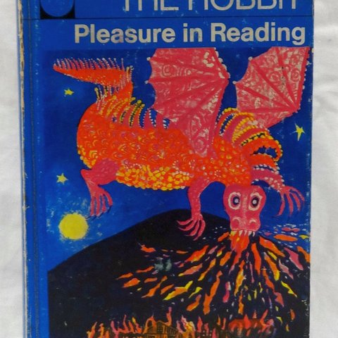 A worn blue paperback copy of The Hobbit with the words “Pleasure in Reading” across the top, featuring a bright red dragon with spread wings breathing fire over an erupting volcano and burning village, and the author credit “J R R Tolkien” in white text