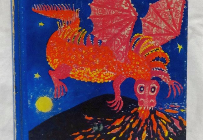 A worn blue paperback copy of The Hobbit with the words “Pleasure in Reading” across the top, featuring a bright red dragon with spread wings breathing fire over an erupting volcano and burning village, and the author credit “J R R Tolkien” in white text