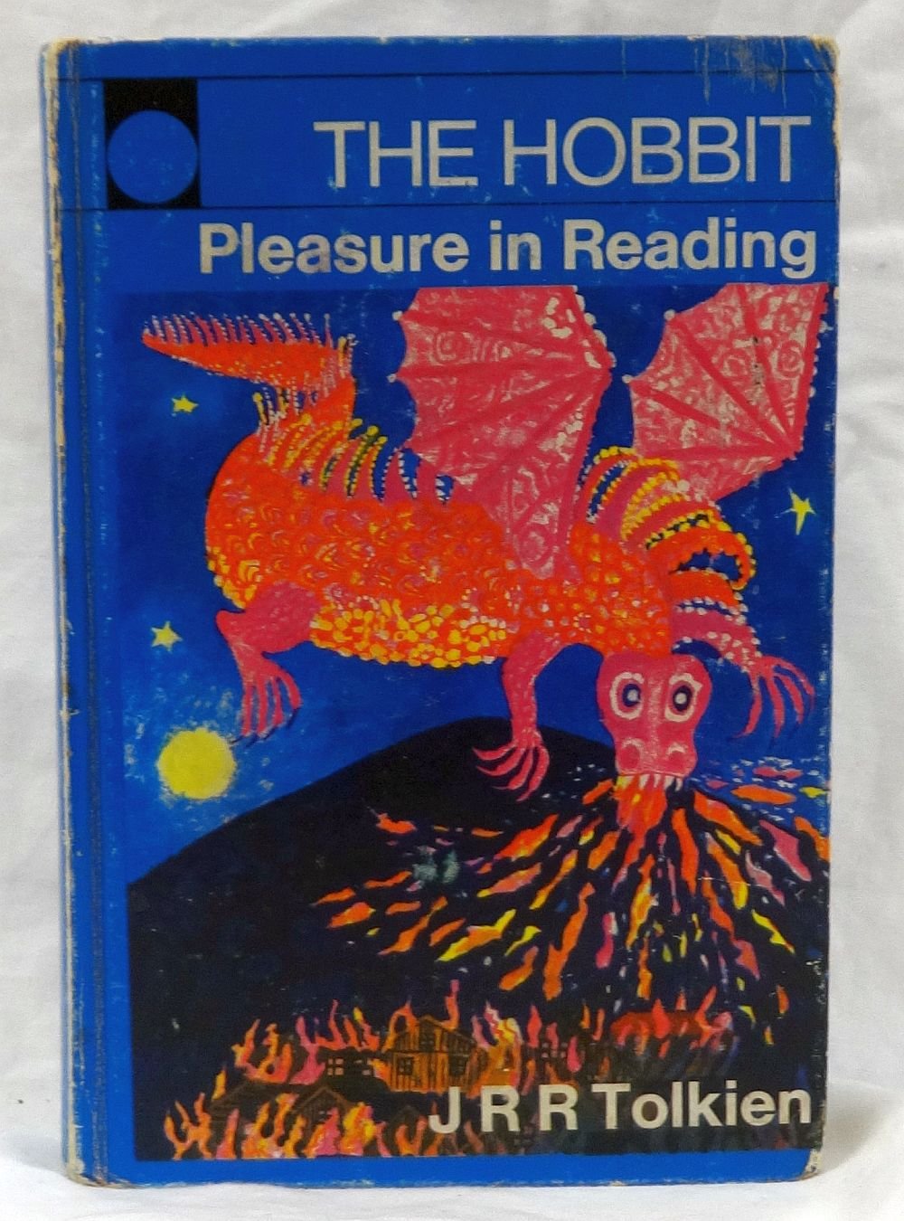 A worn blue paperback copy of The Hobbit with the words “Pleasure in Reading” across the top, featuring a bright red dragon with spread wings breathing fire over an erupting volcano and burning village, and the author credit “J R R Tolkien” in white text