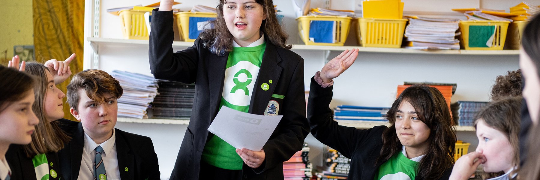 School kids in green Oxfam t-shirts raise their hands.