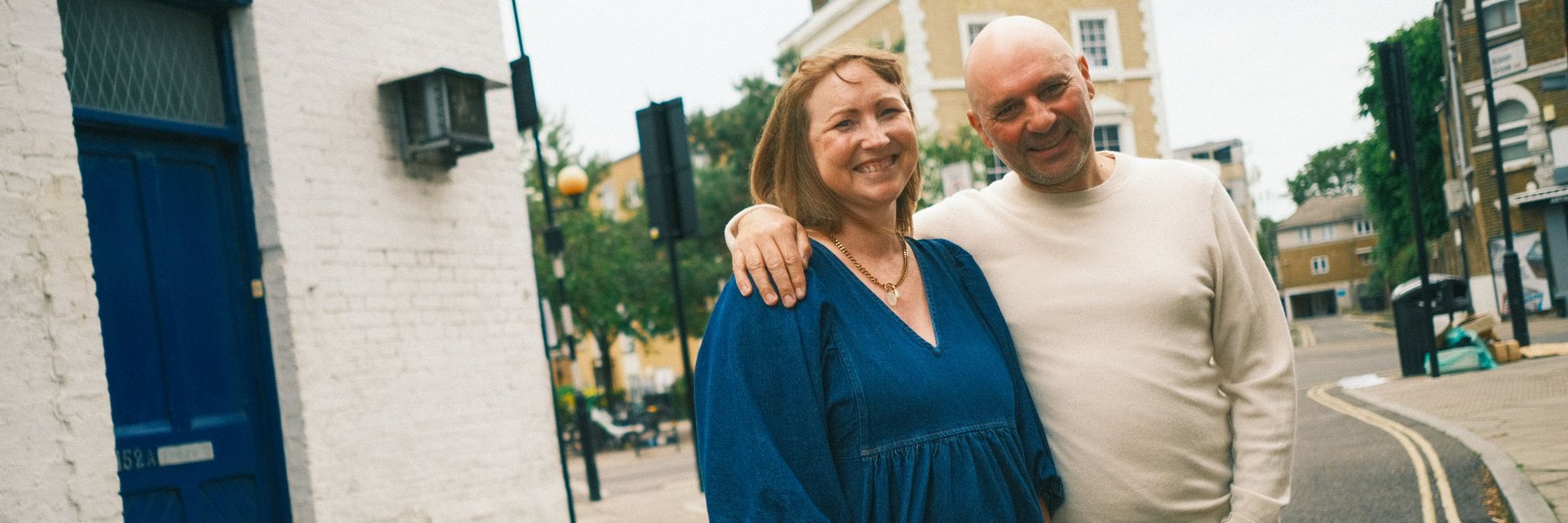 A smiling couple stands arm in arm on a quiet street corner, with brick buildings and a blue door in the background. The woman is wearing a blue dress, and the man is dressed in a light sweater and jeans.