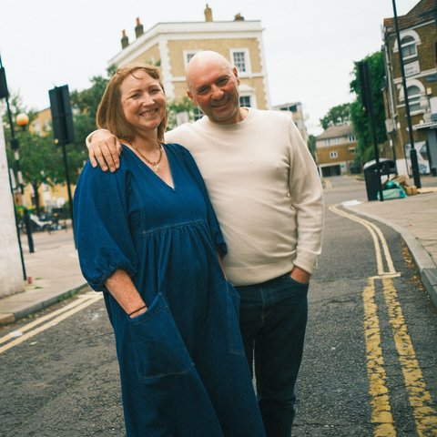 A smiling couple stands arm in arm on a quiet street corner, with brick buildings and a blue door in the background. The woman is wearing a blue dress, and the man is dressed in a light sweater and jeans.