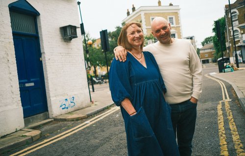 A smiling couple stands arm in arm on a quiet street corner, with brick buildings and a blue door in the background. The woman is wearing a blue dress, and the man is dressed in a light sweater and jeans.