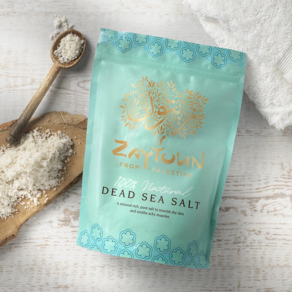 Zaytoun's dead sea salt blue cosmetic pouch on a white table next to sea salt on a spoon