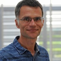 A man wearing glasses and a blue denim shirt smiles into the camera.