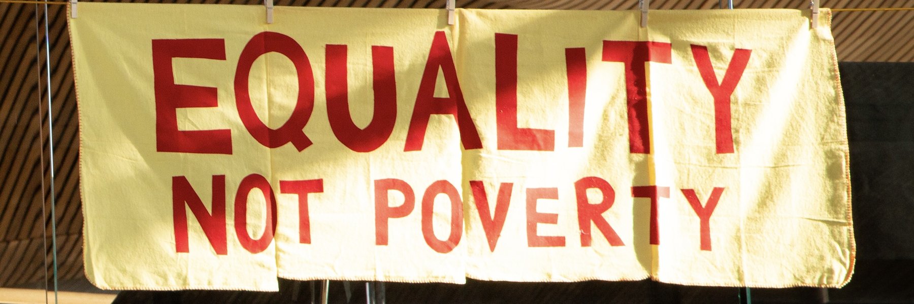An embroidered craftivist banner made from large yellow household dusters hang in the Welsh Senedd, showing the message ‘Equality not Poverty’.