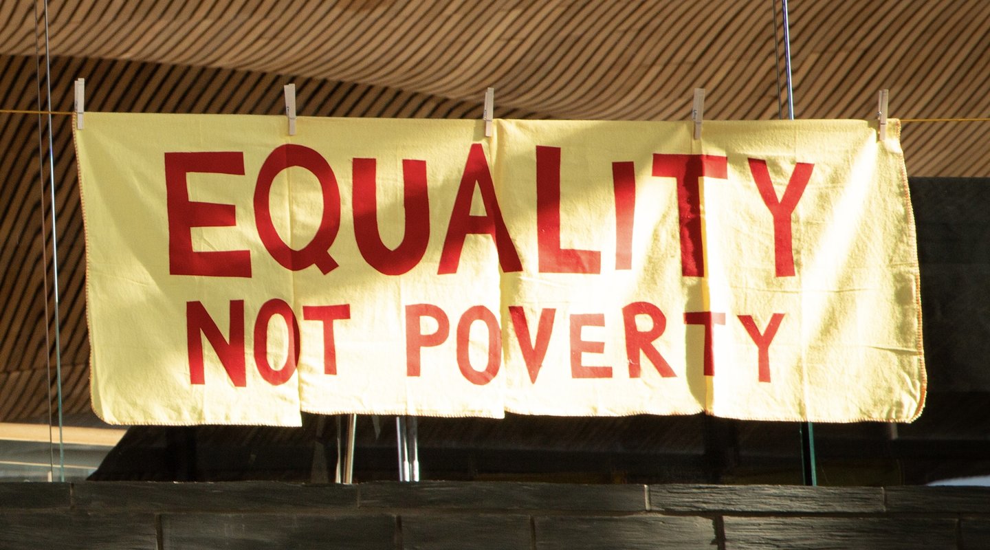 An embroidered craftivist banner made from large yellow household dusters hang in the Welsh Senedd, showing the message ‘Equality not Poverty’.