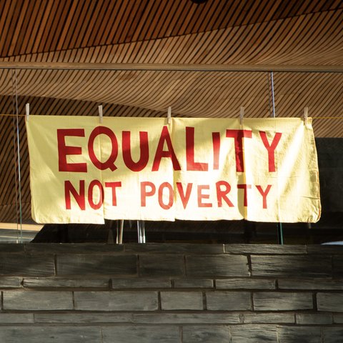 An embroidered craftivist banner made from large yellow household dusters hang in the Welsh Senedd, showing the message ‘Equality not Poverty’.