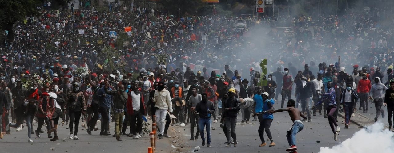 Hundreds people in a street at a protest in Kenya. Some people are throwing objects, some are running and there is smoke rising from the ground.