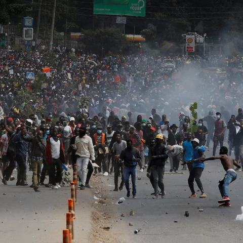 Hundreds people in a street at a protest in Kenya. Some people are throwing objects, some are running and there is smoke rising from the ground.