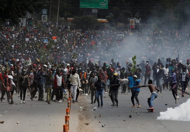 Hundreds people in a street at a protest in Kenya. Some people are throwing objects, some are running and there is smoke rising from the ground.