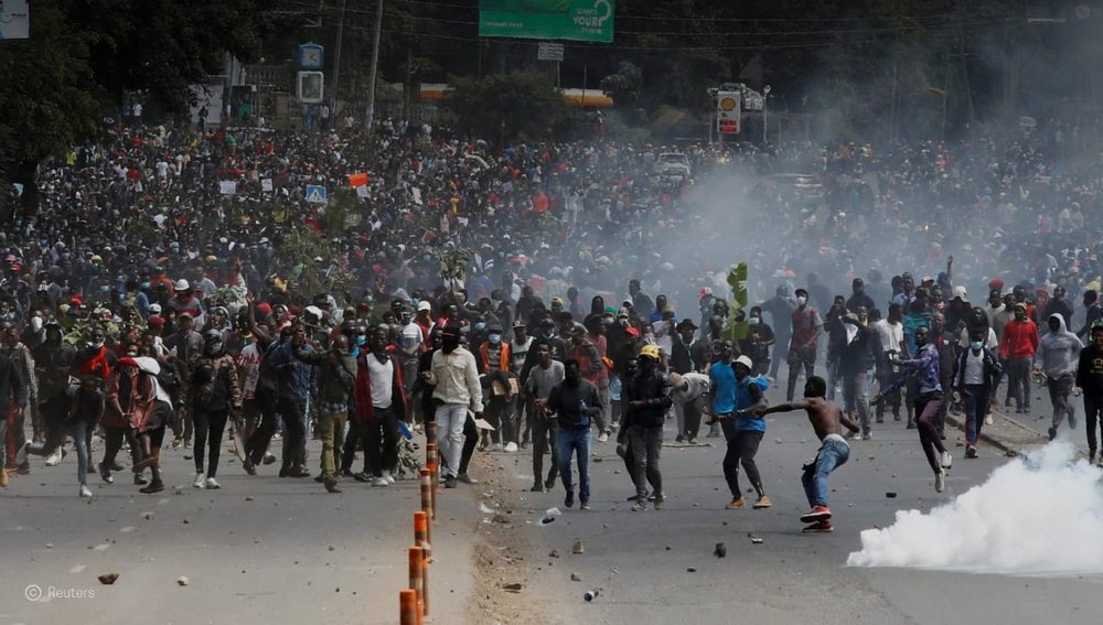 Hundreds people in a street at a protest in Kenya. Some people are throwing objects, some are running and there is smoke rising from the ground.