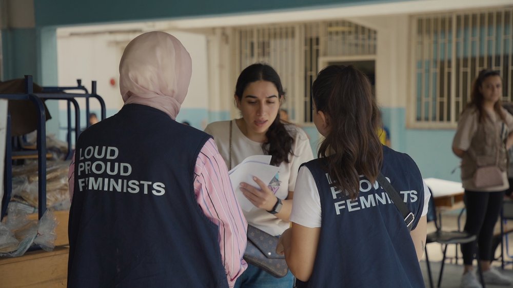 Two women wearing vests that read "LOUD PROUD FEMINISTS" speak with another woman holding papers at a community setting indoors.