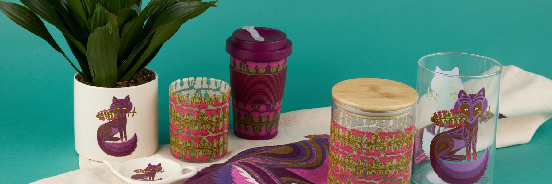 A collection of decorative household items featuring a purple fox design, including a plant pot, travel mug, glass jars, and textiles, displayed against a teal background.