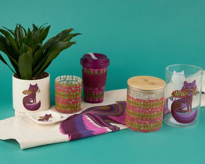 A collection of decorative household items featuring a purple fox design, including a plant pot, travel mug, glass jars, and textiles, displayed against a teal background.