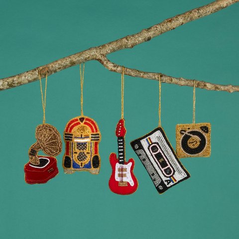Five beaded ornaments shaped like a gramophone, jukebox, electric guitar, cassette tape, and camera hanging from a branch against a green background.