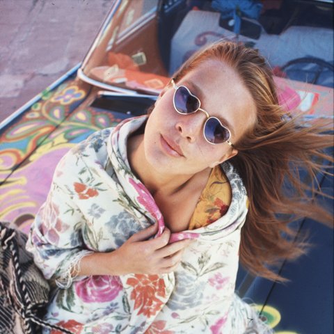 A woman wearing heart-shaped sunglasses, with a flower-printed shawl wrapped around her. She sits on a car bonnet that is spray painted with psychedelic patterns.