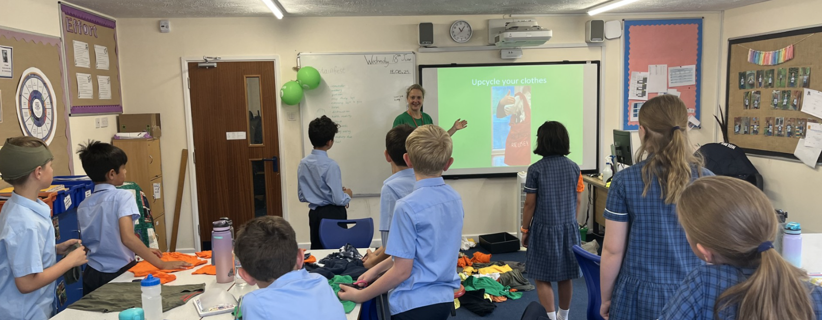 A classroom with children in school uniforms gathered around tables covered with fabric pieces. At the front, a person stands near a screen displaying a slide titled ‘Upgrade your clothes.’