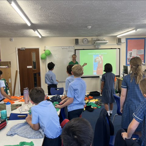 A classroom with children in school uniforms gathered around tables covered with fabric pieces. At the front, a person stands near a screen displaying a slide titled ‘Upgrade your clothes.’
