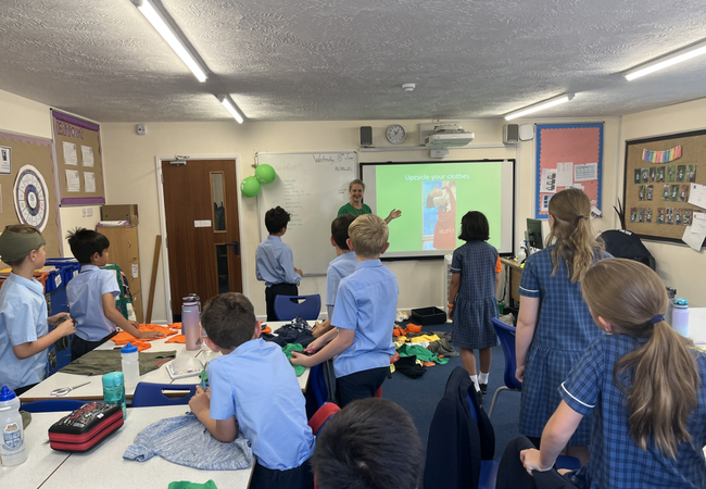 A classroom with children in school uniforms gathered around tables covered with fabric pieces. At the front, a person stands near a screen displaying a slide titled ‘Upgrade your clothes.’