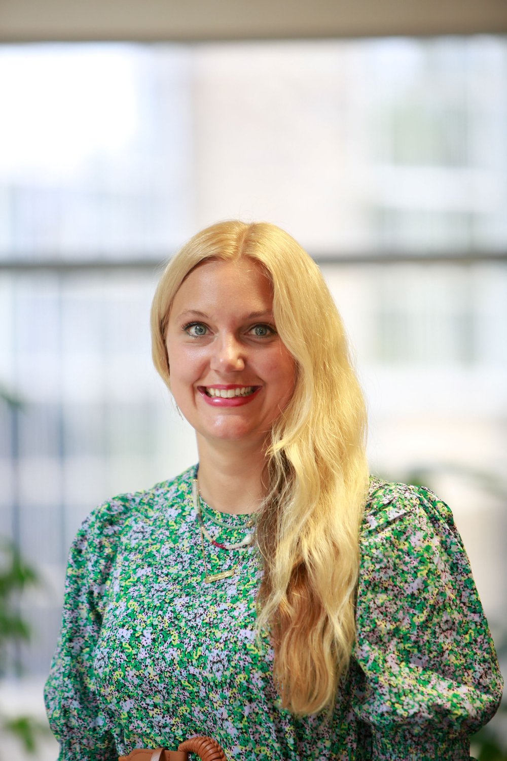 A woman with long blonde hair smiling, wearing a green floral top, standing indoors with soft natural light in the background.
