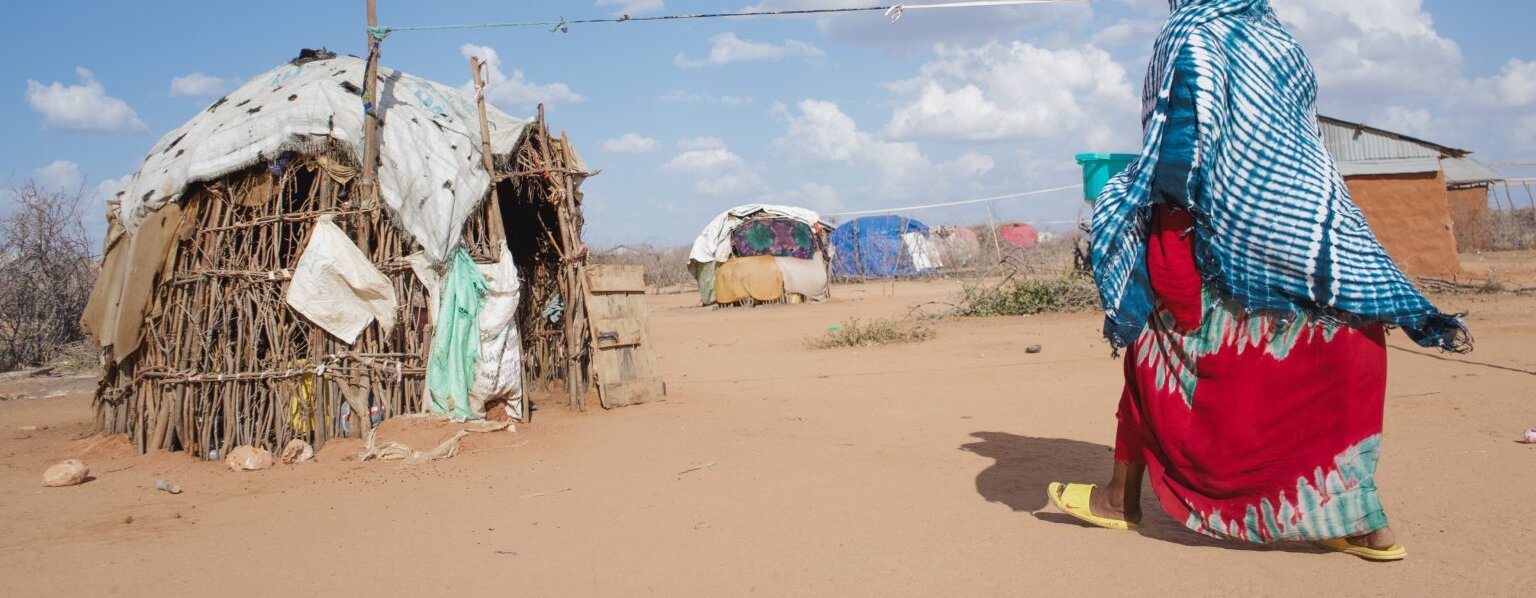 Hadija dressed in brightly coloured clothes walking towards wooden hut on sandy ground with blue sky.