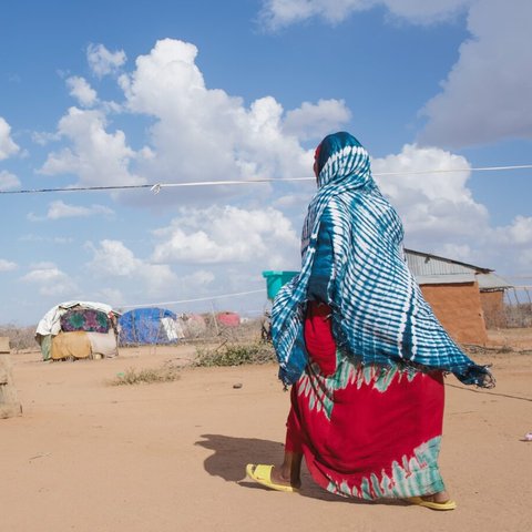 Hadija dressed in brightly coloured clothes walking towards wooden hut on sandy ground with blue sky.