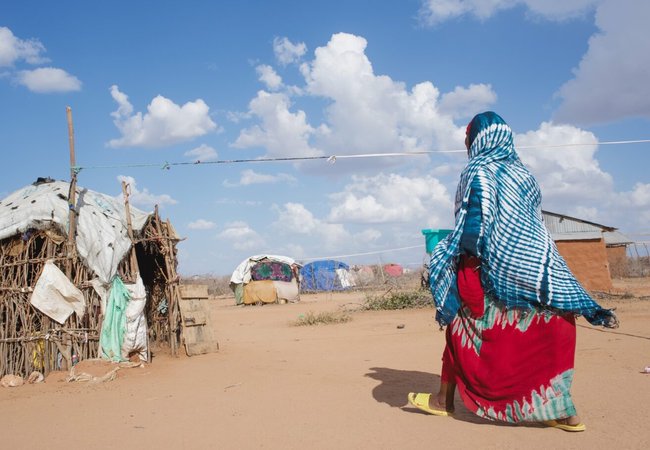 Hadija dressed in brightly coloured clothes walking towards wooden hut on sandy ground with blue sky.