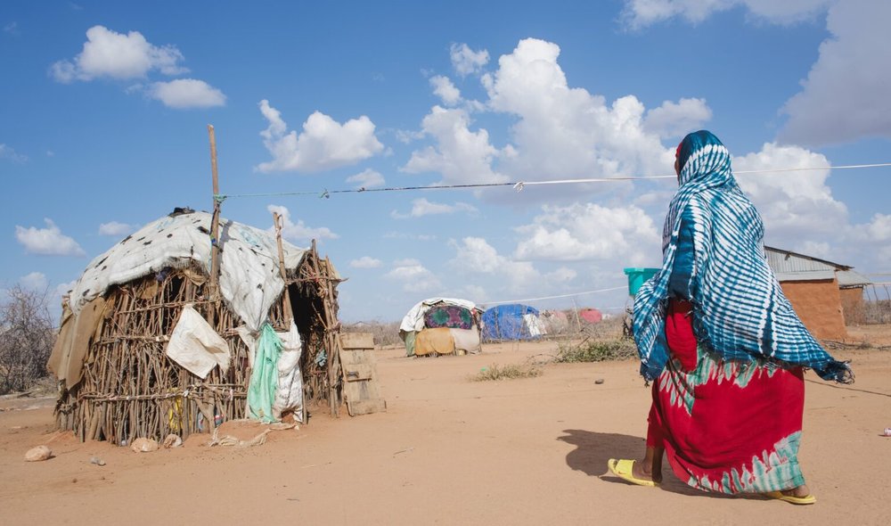 Hadija dressed in brightly coloured clothes walking towards wooden hut on sandy ground with blue sky.