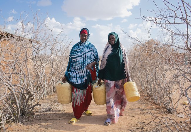 Hadija Jillo and another woman walking to fill up yellow jerry cans on parched ground