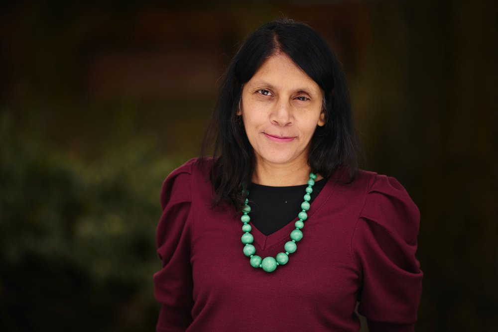 A woman with black hair in a burgundy top and green necklace, smiling softly outdoors.