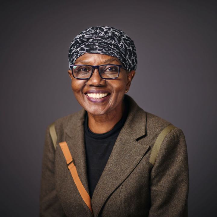 A woman in glasses smiles warmly at the camera. She is wearing a leopard print head covering, a black t-shirt and a brown blazer. The background is a plain dark grey.