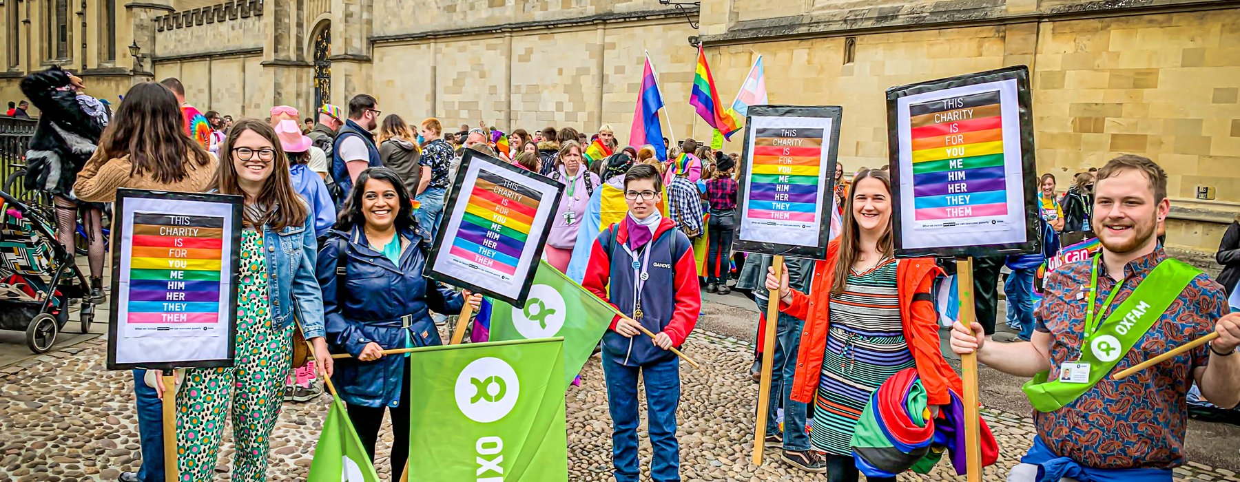 5 people hold Oxfam Pride Posters and Oxfam flags standing on cobbles with an Oxford University college behind them.