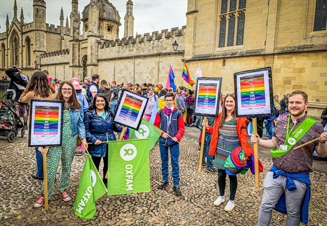5 people hold Oxfam Pride Posters and Oxfam flags standing on cobbles with an Oxford University college behind them.