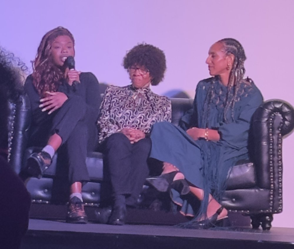 Three people sit on a black sofa on-stage at the Black Writers Guild Conference. Busayo Twins is holding a microphone and speaking to the audience.