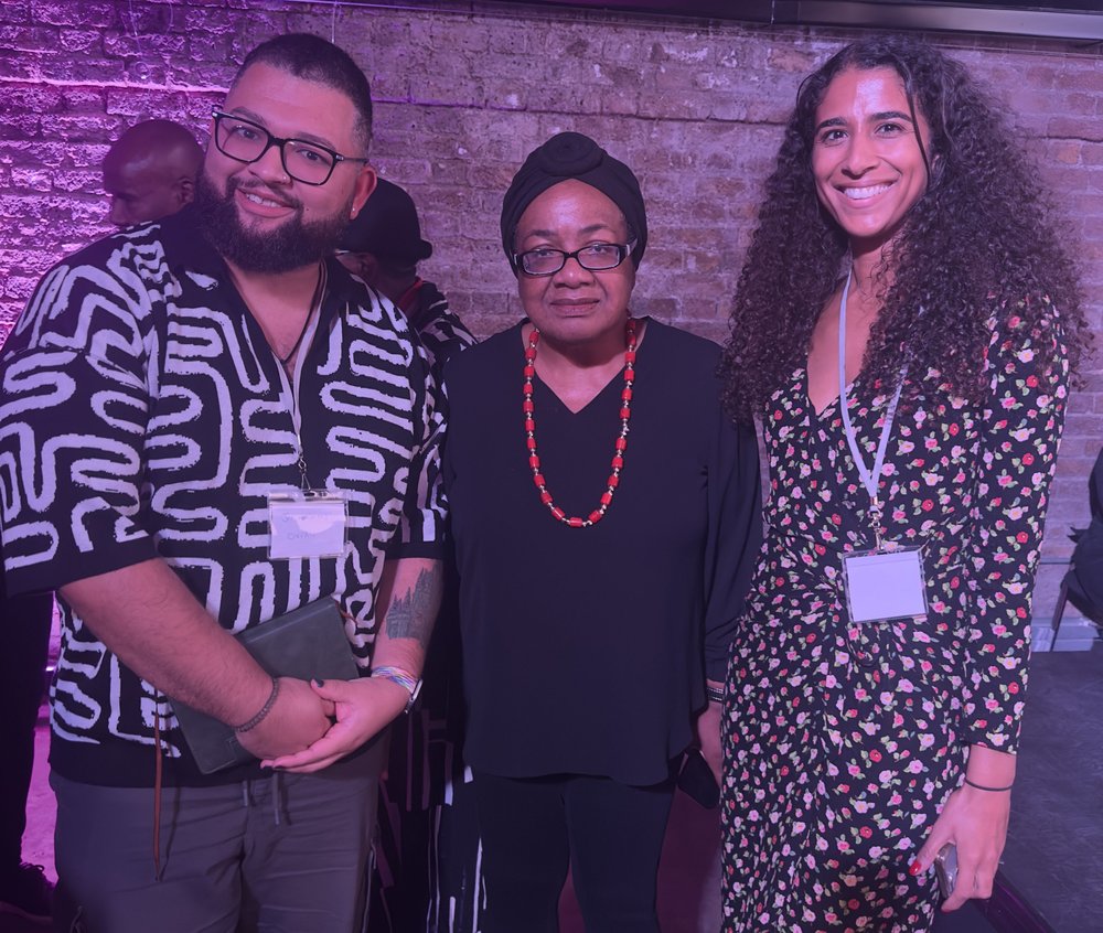 Three people pose together and smile at the Black Writers Guild conference, standing in front of a brick wall with purple lighting.