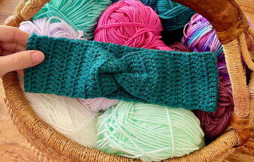 A green crochet headband is resting inside a wicket basket on top of different coloured balls of yarn.