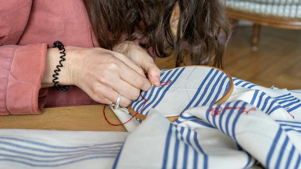 A person using a embroidery hoop and red thread to mend a blue and white striped piece of clothing.